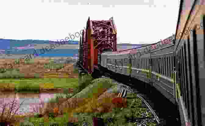 A Photograph Of A Trans Siberian Railway Train Crossing A Bridge Over A River In Siberia. The Train Is Painted In White And Green, And It Is Dwarfed By The Vast Landscape Around It. The End Of The Line